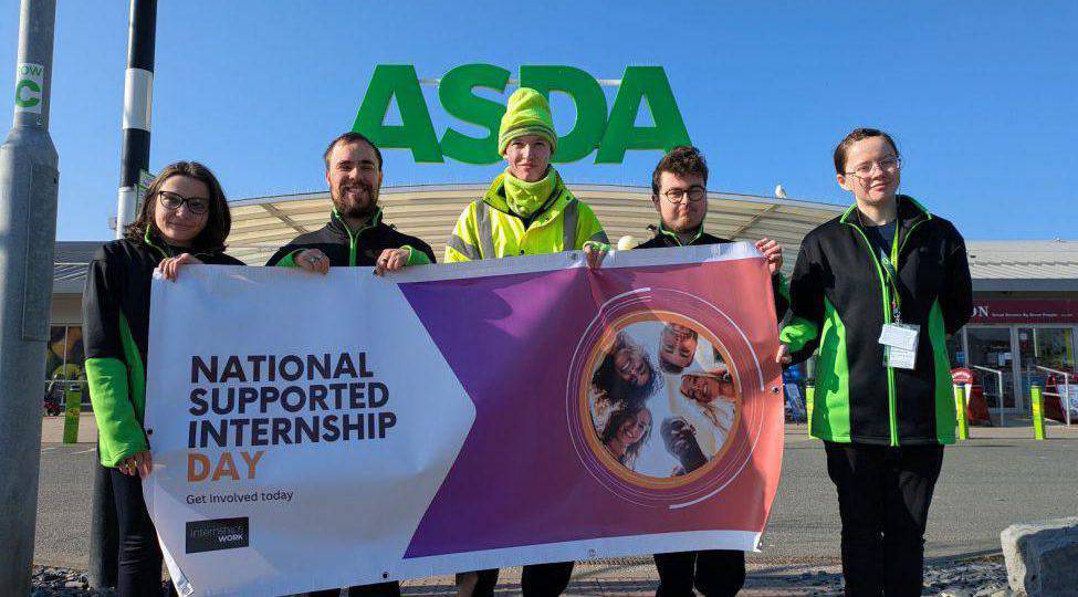 Dysgwyr y rhaglen Interniaeth a Gefnogir yn Asda Llandudno yn gyda baner yn hyrwyddo Diwrnod Cenedlaethol Interniaethau a Gefnogir